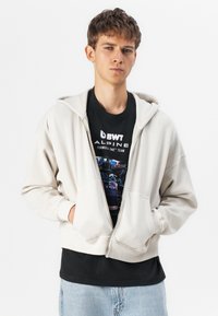 Haut à capuche zippé beige clair en mélange de coton, doté d'une fermeture éclair à l'avant, de deux poches et de poignets côtelés. Porté par-dessus un t-shirt graphique noir.