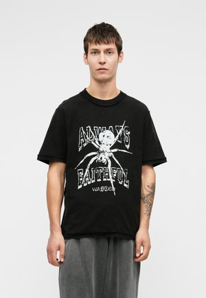 Fiatal férfi áll, fekete pólót visel, rajta egy nagy fehér pókháló grafika és a "Mindig Hű Elpazarolt" felirat, szürke nadrágot hord, és a bal alkarján tetoválás látható.