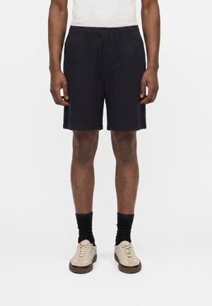 Férfi fekete zsinóros rövidnadrágban, fekete zokniban, bézs sportcipőben és fehér ingben, fehér háttér előtt áll.