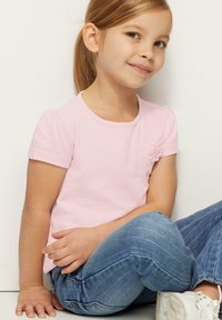 Helles rosa T-Shirt mit kurzen Ärmeln, gerafften Ärmeln und einem floralen Akzent an der Schulter, kombiniert mit blauen Jeans und weißen Schuhen.