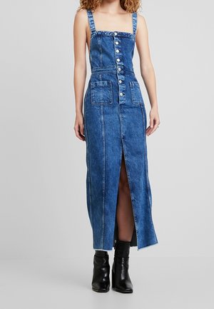 Femme portant une robe midi en denim bleu sans manches avec des boutons, des poches à l'avant et une fente devant, associée à des bottines noires.
