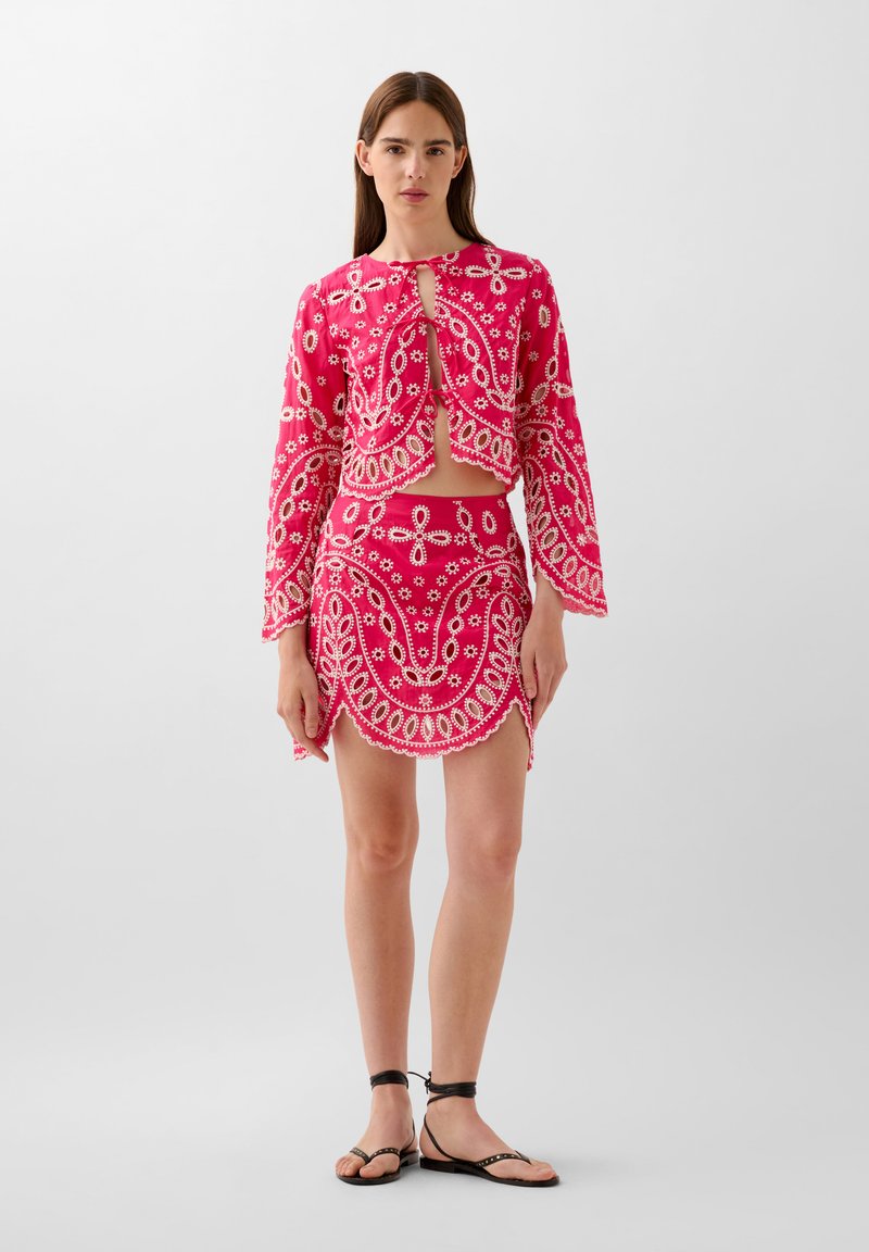 Un top court rose vif et une jupe assortie avec des détails de broderie florale et paisley blancs. La jupe présente un ourlet courbé et une fente sur le côté.