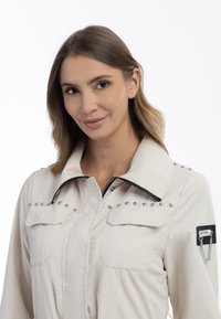 Jachetă crem deschis cu fermoar, având accente din studs metalici pe guler și buzunare, cu un patch negru și detaliu din lanț pe mânecă.