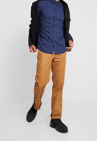 Chemise bleu marine à boutons avec des pois blancs, associée à un pantalon beige et des chaussures noires. La chemise possède un col classique et des manches longues.