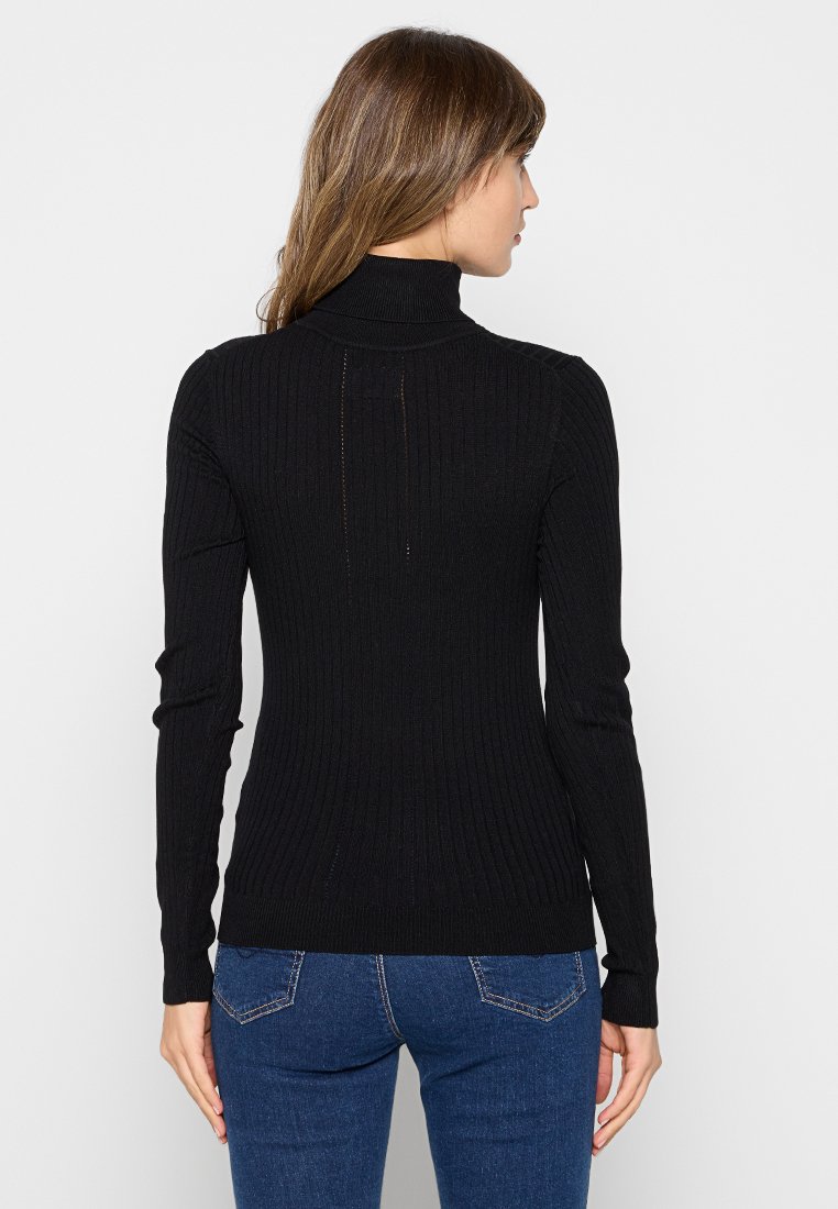 Pull à col roulé noir côtelé avec des manches longues, présentant un design ajusté et un détail discret au dos. Associé à un jean bleu.