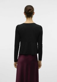 Svart långärmad topp med kroppsnära midja och utsvängd nederdel. Mjuk textur och scallopad halsringning. Bärs över en bordeauxröd kjol.