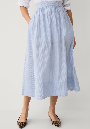 Kobieta ubrana w niebiesko-białą, pionowo paskowaną spódnicę midi z kieszeniami, zestawioną z płaskimi butami na obcasie typu slingback w panterkę o spiczastym nosku.