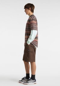 Camisa de manga corta a rayas en colores vibrantes, combinada con pantalones cortos cargo marrones y zapatillas grises, con un acento azul y logo.