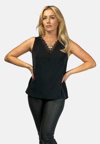 Czarny bezrękawnik z falbaniastym koronkowym dekoltem, gładki materiał i luźny krój, w połączeniu z czarnymi dopasowanymi spodniami.