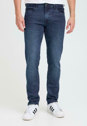 Dark blue denim jeans, slim fit, with five pockets and a subtle fade. Worn with white sneakers featuring black stripes.
