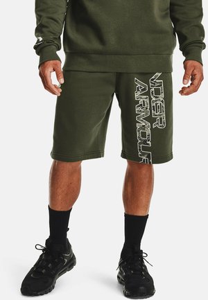 Homme portant un short Under Armour vert olive avec un grand logo blanc sur la jambe gauche, des chaussettes noires et des chaussures de sport noires.