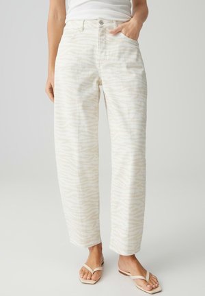 Vrouw draagt hoge taille, loszittende crèmekleurige broek met subtiele zebrastripes, wit ingestopt shirt en beige sandalen, staand op een lichte achtergrond.