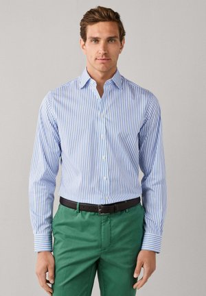 Man draagt een blauw-wit gestreept overhemd met knopen, ingestopt in groene broek met een zwarte riem, staand tegen een effen achtergrond.