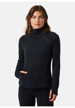 Donna che indossa una giacca nera con cerniera, colletto alto e fori per il pollice, in piedi davanti a uno sfondo chiaro uniforme, con una mano in tasca.