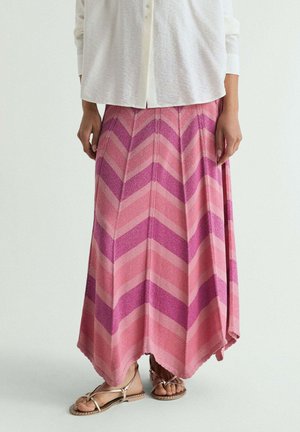 Mujer con falda de patrón Chevron en rosa y púrpura, blusa blanca con botones y sandalias beige con tiras, de pie sobre un fondo blanco.
