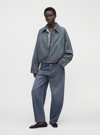 Personne portant une veste en jean grise ample et un jean large avec des chaussures plates noires, debout devant un fond clair uni.
