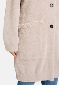 Jasnoróżowy futrzany płaszcz o fakturowanej powierzchni, duża boczna kieszeń z frędzlami oraz cztery czarne guziki z przodu.