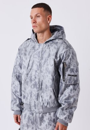 Man draagt een grijze bomberjack met patroon en capuchon met een ritssluiting op de mouw en bijpassende broek, kijkt naar links tegen een effen achtergrond.
