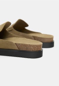 Șlapi din suede maro cu talpă intermediară din plută și talpă exterioară din cauciuc negru, având o textură netedă și un design simplu.