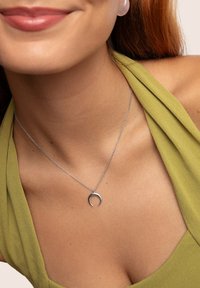 Collana d'argento con pendente a mezzaluna minimalista, indossata su un top di stoffa verde con una trama morbida e drappeggiata.