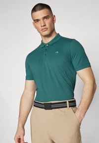 Tirkizna polo majica s kratkim rukavima, gumbima i CK logotipom. Nosena s bež hlačama i crnim remenom s prugastim detaljima.