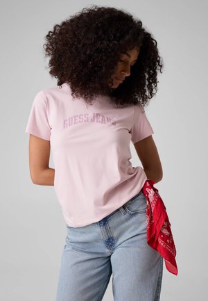 Frau mit lockigem Haar, die ein pinkes Guess Jeans T-Shirt und hellblaue Jeans trägt, aus deren Tasche ein rotes Bandana hängt.