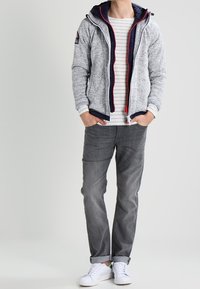 Man med grå zip-up hoodie över randig vit skjorta, mörkgrå jeans och vita sneakers med händerna i fickorna.