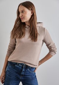 Felpa beige realizzata in tessuto morbido, con tasca frontale e polsini a costine, abbinata a jeans blu. Il modello si trova in una posizione rilassata.