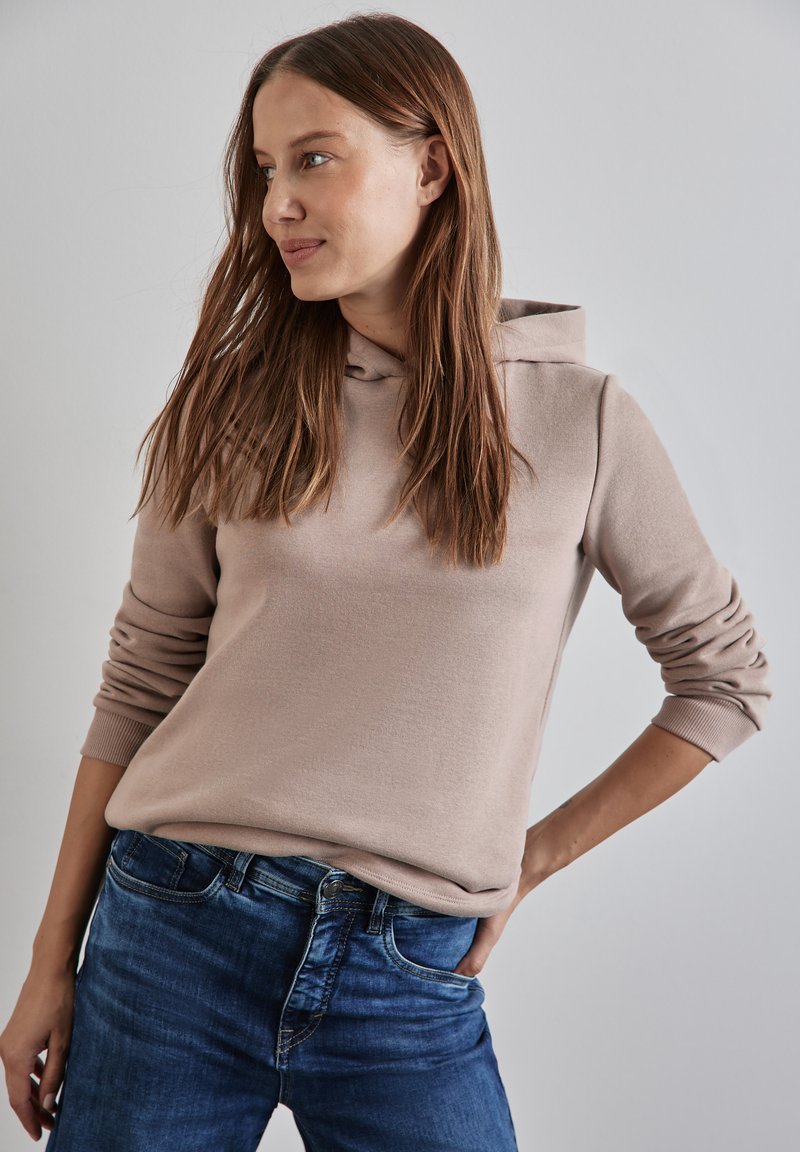 Felpa beige realizzata in tessuto morbido, con tasca frontale e polsini a costine, abbinata a jeans blu. Il modello si trova in una posizione rilassata.