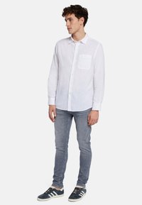 Chemise en lin blanche à manches longues avec col boutonné et poche poitrine unique, associée à un jean slim bleu clair et des baskets sombres.