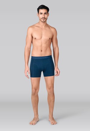 Bóxers tipo briefs en azul marino oscuro, hechos de material suave, con una cintura elástica lisa que lleva el logo "MUCHACHO MALO" y detalles de costuras sutiles.