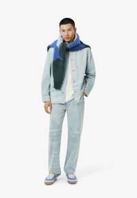 Lichtblauwe denimjack over een wit shirt, gecombineerd met lichtblauwe jeans met een subtiele print. Een kleurrijke, harige sjaal hangt om de hals.