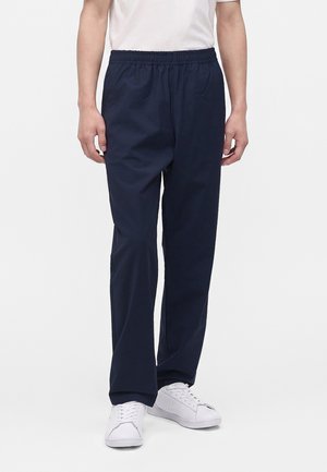 Personne portant un pantalon bleu marine à taille élastique et des baskets blanches, debout devant un fond clair uni.