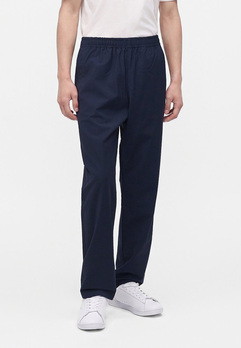 Personne portant un pantalon bleu marine à taille élastique et des baskets blanches, debout devant un fond clair uni.