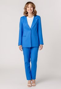 Blue suit set made of textured fabric, featuring a tailored blazer with lapels and straight-leg trousers, paired with a white top and heels.