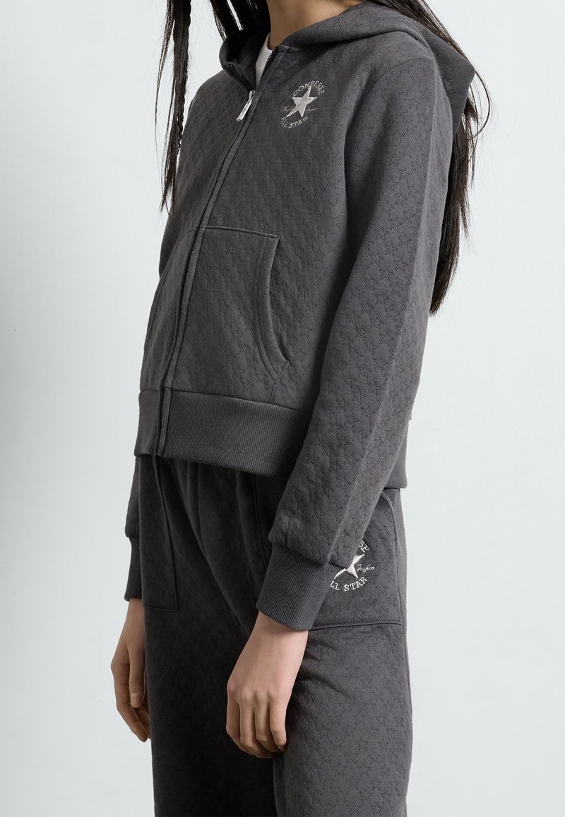 Jeune personne portant un sweat à capuche matelassé gris foncé zippé et un pantalon assorti avec les logos Converse All Star sur la poitrine et la cuisse.