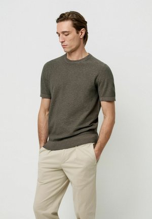 Maglione a maniche corte, verde oliva scuro, lavorato a maglia con scollo rotondo; abbinato a pantaloni beige, che evidenziano una texture liscia e linee pulite.