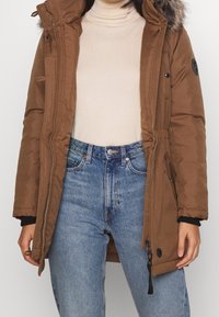 Brown parka jacket with a faux fur hood, zip closure, adjustable waist, and side pockets, paired with light blue jeans and a beige top.
