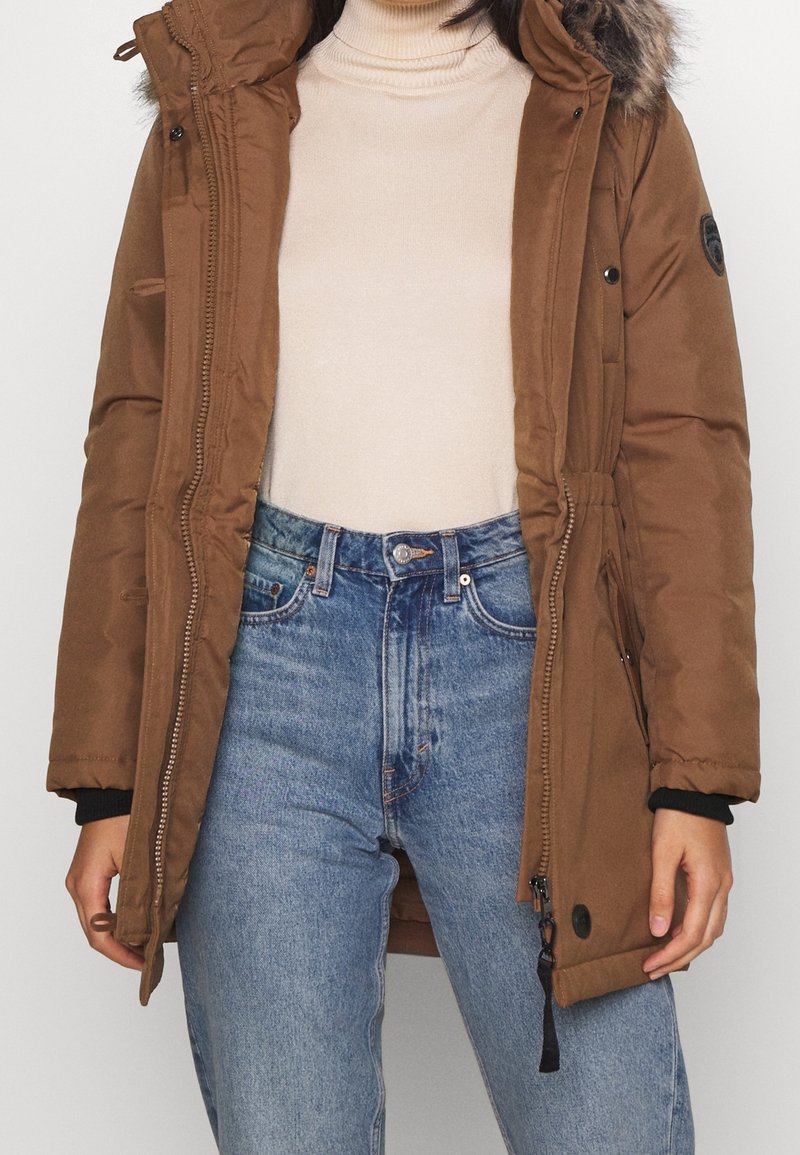 Brown parka jacket with a faux fur hood, zip closure, adjustable waist, and side pockets, paired with light blue jeans and a beige top.