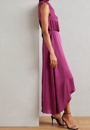 Frau trägt ein ärmelloses magentafarbenes Midikleid mit asymmetrischem Saum und schwarzen High-Heel-Sandalen, steht seitlich an einer Wand.