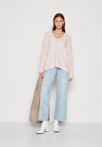 Lichtroze, losgebreide trui met V-hals, gecombineerd met lichtblauwe wijde jeans. Het model draagt witte enkellaarsjes en houdt een beige jas vast.
