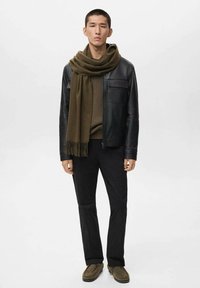 Giubbotto di pelle nera con zip, sciarpa verde oliva, maglione a coste, pantaloni neri e scarpe grigie. Modello in piedi contro uno sfondo neutro.