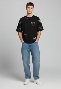 Black short-sleeve T-shirt with embroidered designs, including hearts, butterfly, and text; paired with light blue jeans and white sneakers.