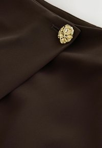 Dunkelbrauner Stoff mit einem strukturierten goldenen Knopf, der durch ein genähtes Knopfloch an einem Kleidungsstück befestigt ist.