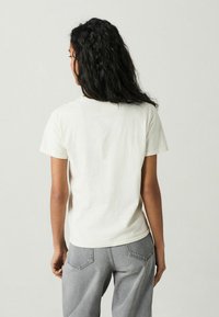 Weißes Baumwoll-T-Shirt mit kurzen Ärmeln und Rundhalsausschnitt, von hinten betrachtet; kombiniert mit hellgrauen Hosen mit Gesäßtaschen.