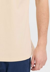 Lys beige polo skjorte med en strukturert overflate, med en subtilt buet kant og en liten merkeetikett på side sømmen. Mørkeblå shorts synlige.