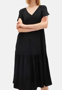 Žena v volných černých šatech s krátkými rukávy a výstřihem do V, se zkříženýma rukama, stojící na jednolitém světlém pozadí.