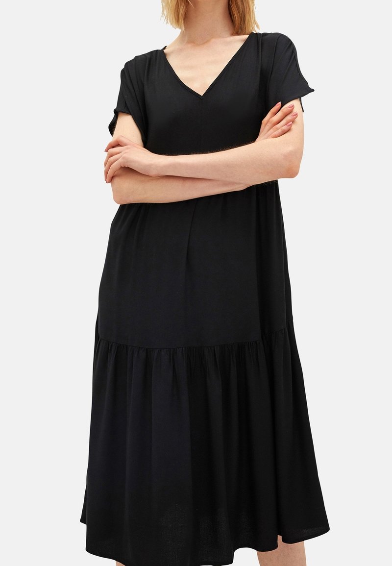 Žena v volných černých šatech s krátkými rukávy a výstřihem do V, se zkříženýma rukama, stojící na jednolitém světlém pozadí.