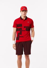 Röd polo skjorta med kontrasterande svarta geometriska mönster, svart krage och Lacoste-logotyp. Matchad med mörkröda shorts och en passande keps.