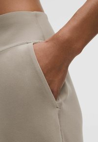 Eine Hand ruht in der Tasche von hellbeigen, hoch geschnittenen, nahtlosen, enganliegenden Hosen vor einem schlichten Hintergrund.